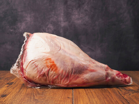 Raw lamb leg in plastic vacuum wrap rests on wooden surface, painted background. Meat shows pink flesh and red fat marbling. Scene suggests butchery, cooking, or grocery retail.