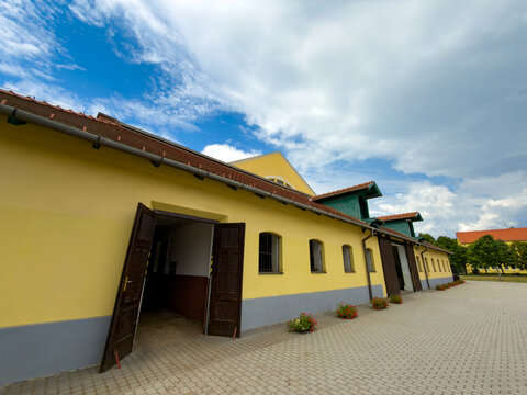 Exterior view of yellow barn building with open doors. Red potted flowers line a stone paved yard under a blue sky with white clouds during daytime.
