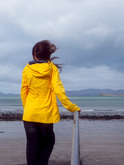 Person in bright yellow jacket stands on stone steps by coastal path. They face calm sea under cloudy sky. View shows distant shoreline and rocky beach © mark_gusev
