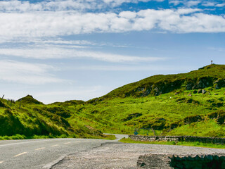 Fototapeta premium Winding road cuts through lush green hills under bright blue sky with fluffy clouds. Grassy slopes rise gently, dotted with wildflowers and scattered stones. Small stone wall borders path