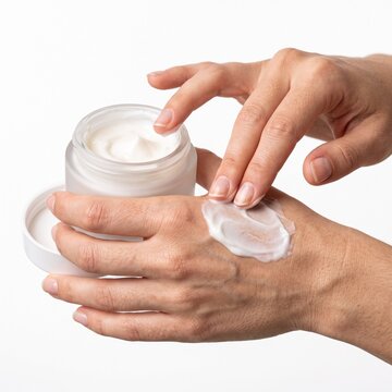 A close-up shot of a person applying a white cream from a glass jar onto their hand. The person's fingers are gently massaging the cream into their skin, showing a moment of self-care and hydration
