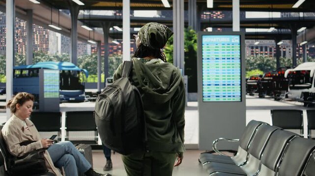 Female traveler walks through a bus station with coffee and luggage, carrying a takeaway drink and backpack into the coach transit waiting area.