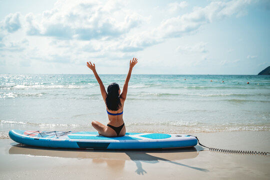 Fit Asian woman sits on a SUP, arms up, experiencing liberation and joy during a beach retreat