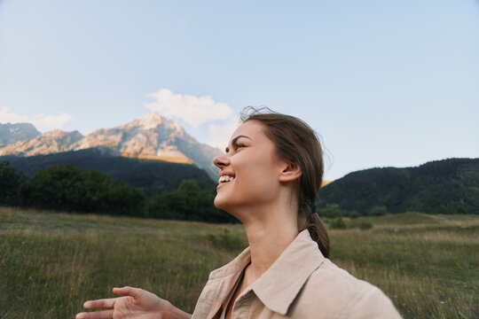 A smiling woman with braided hair enjoys a sunny day outdoors in a grassy field with mountains in the background, capturing a joyful lifestyle moment in nature.