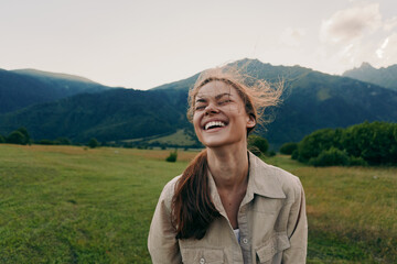 Fototapeta premium A smiling young woman stands in a green meadow with mountains in the background, wind in her hair, capturing a carefree outdoor moment, with light suggesting freedom today.