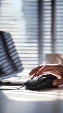 Person using a wired black computer mouse with sunlight streaming through window blinds onto a minimalist white desk and keyboard in office setting