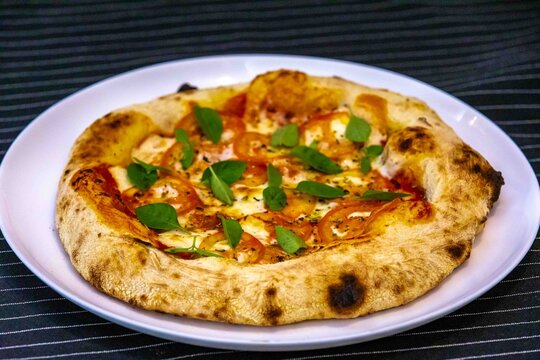 Neapolitan-style personal pizza with naturally leavened long-fermentation dough, sliced tomatoes, melted mozzarella, fresh basil and oregano, served on a white plate over a dark pinstriped cloth.