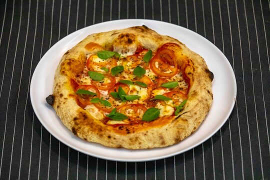Neapolitan-style personal pizza with naturally leavened long-fermentation dough, sliced tomatoes, melted mozzarella, fresh basil and oregano, served on a white plate over a dark pinstriped cloth.