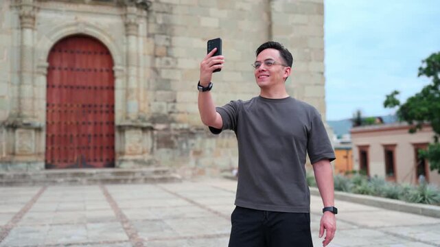 Young man smiling and taking a selfie video with his smartphone while visiting the historic center of oaxaca, mexico, enjoying his vacation and sharing his travel experience online