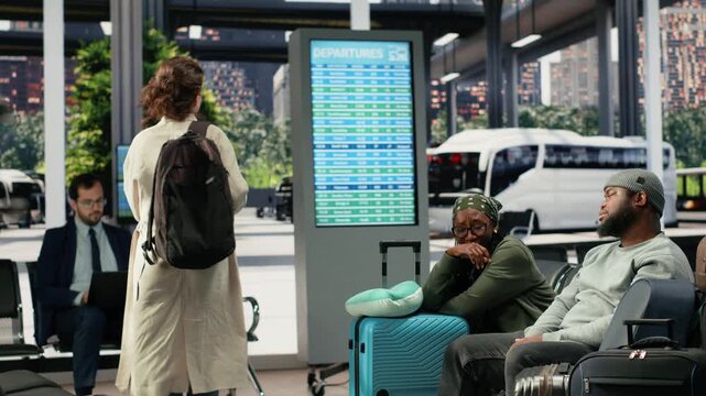 Tired tourists sit on a bus terminal bench and discuss the delay, with an African American couple talking beside other passengers while waiting for departure.
