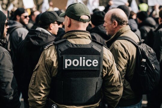 Police officer in bulletproof vest stands in crowd at public protest. Man wearing cap and jacket watches demonstration. Law enforcement works public order.