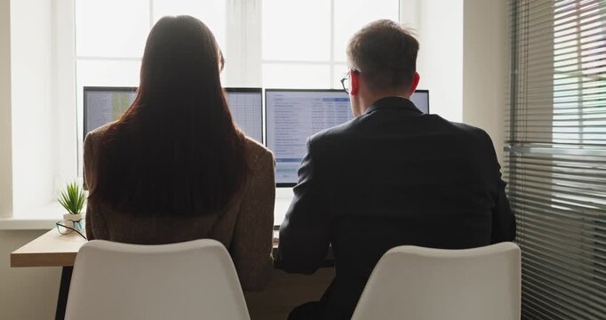 Business colleagues teamwork on financial analysis today. Rear view of man and woman at dual computer monitors, reviewing data. Focus on budgeting and reporting. Clear concept of business teamwork.