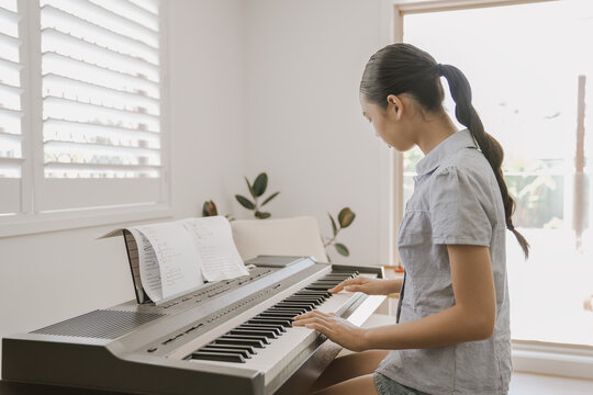 piano, music, woman, home, playing, keyboard, practicing - A young Asian woman focuses on practicing a song on her digital piano in a bright living room, capturing a peaceful moment.