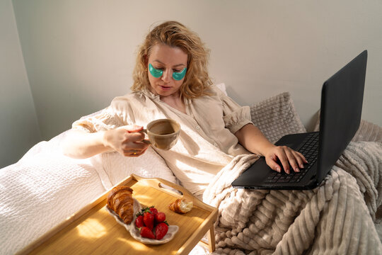 A blonde woman working in bed with green eye patches, typing on her laptop while holding a cup of coffee next to a breakfast tray with croissants and strawberries