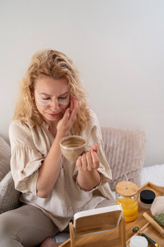 Beautiful natural woman in early forties drinks coffee in her bed while having beauty morning routine before work 
