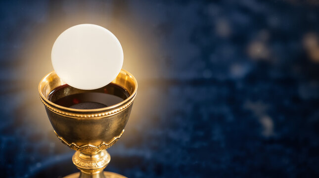 Holy communion wafer resting above a golden chalice of red wine. Christian sacrament of the Eucharist with divine golden glow. Dark blue background with copy space