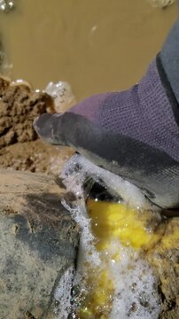 Vertical close-up of a utility worker detecting a dangerous natural gas pipeline leak using a soap bubble test in a muddy trench.