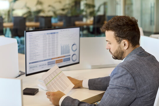 Accountant analyst reviewing report on computer. He checks pages and views charts and spreadsheets in a bright office. Focus on finance data accuracy. Concept financial analysis and reporting.