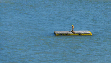 Minimal isolated rectangular wooden floating platform with a yellow edge drifting calmly on open blue water surface under bright natural sunlight, creating a tranquil seascape © John_Doo78