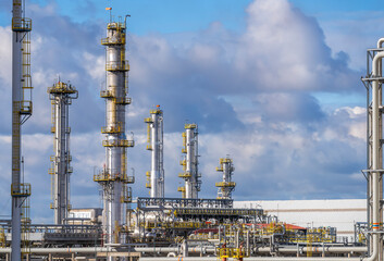 Obraz premium Complex petrochemical refinery plant with tall steel distillation columns, metal pipes, intricate industrial structures under bright blue sky with fluffy white clouds
