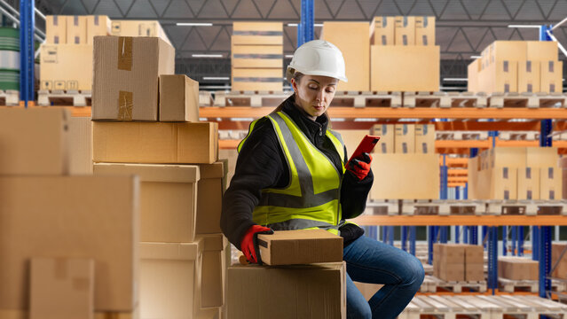 Warehouse logistics. Female warehouse worker checks inventory on a smartphone while handling cardboard packages in a storage and distribution center.