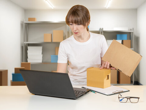 Female small business owner managing e-commerce orders while packing products in home warehouse. Entrepreneur checks inventory and shipping details on laptop while preparing parcels for delivery.