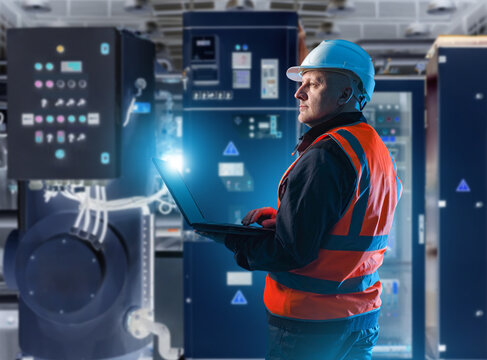 Industrial technician monitoring and controlling heavy machinery systems in a modern automated plant. The scene shows a worker using a laptop to supervise industrial equipment and control panels