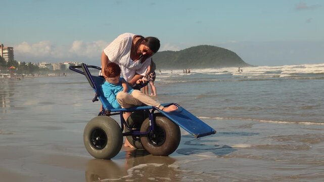 Caregiver helping a child use an all-terrain wheelchair near the ocean, showing accessible beach recreation, inclusion, mobility support and family care.