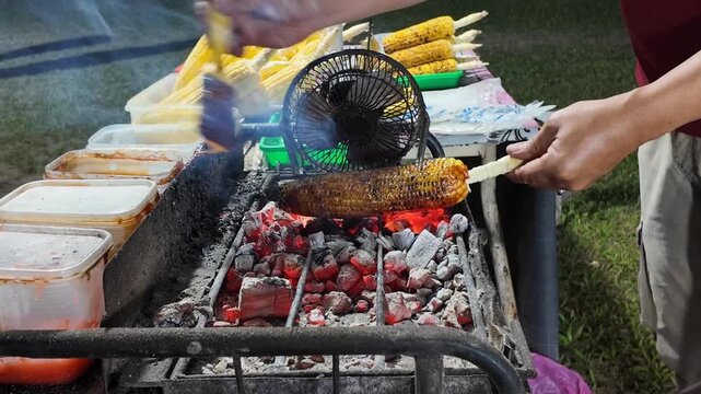 Fresh corn slowly roasts over glowing charcoal while a vendor brushes it with seasoning, filling the fairground air with smoky aromas and creating a tempting street food moment