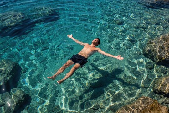 Young man floats relaxed on back in crystal clear Mediterranean Sea water. Male swimmer enjoys summer vacation, feeling free, peaceful. Aerial view of person drifting in turquoise ocean with rocky