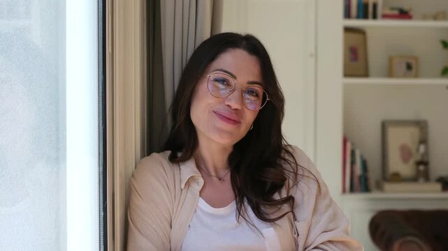 A happy young woman with glasses smiles warmly, looking directly at the camera. She radiates joy and contentment while relaxing comfortably in her home environment near a bright window.