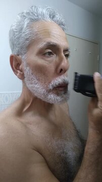 Mature man trimming and shaping his white beard and mustache with an electric clipper