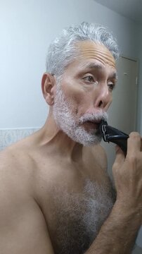 Mature man trimming and shaping his white beard and mustache with an electric clipper