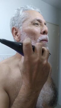 Mature man trimming and shaping his white beard and mustache with an electric clipper