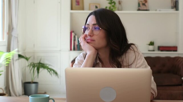 A young woman with glasses sits at her home desk, looking thoughtfully away from her laptop screen. She appears focused or perhaps contemplative while engaged in remote work or study.