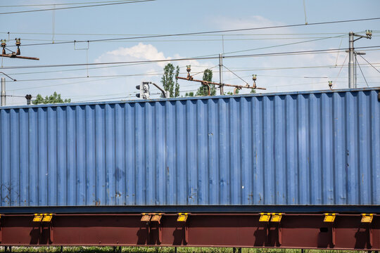 Blue shipping container carried on a cargo train beneath railway power lines, showing intermodal freight transport, rail logistics infrastructure and containerized goods movement across the network.