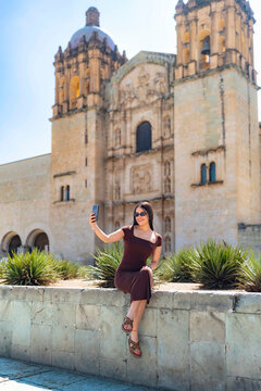 Young woman smiling and taking a selfie on a stone wall in front of santo domingo de guzman church in oaxaca, mexico, enjoying sunny travel vibes and colonial architecture
