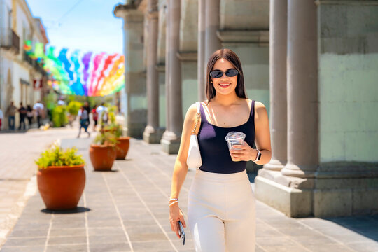 Latina woman smiling, wearing sunglasses, carrying a drink and phone, walking along a sunny street in oaxaca, mexico, with colorful decorations in the background