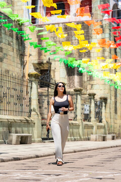 Young latina woman walking along a sunny street in oaxaca, mexico, carrying a drink and phone, with colorful papel picado decorations strung overhead and colonial architecture in the background
