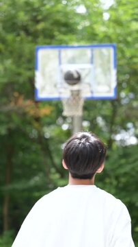 A vertical video showing the back view of a boy shooting a basketball on a lush outdoor court; his face is not visible.
