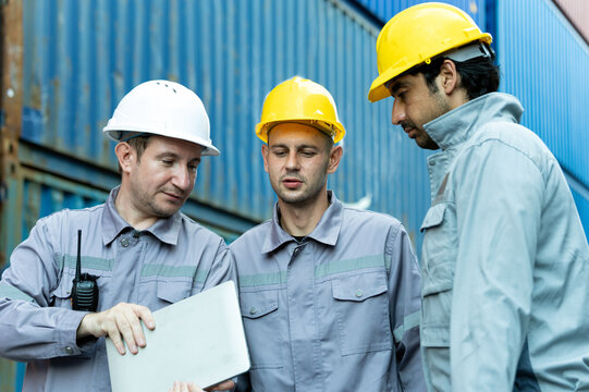 Industrial workers wearing helmets reviewing laptop beside shipping containers, showing logistics, teamwork, safety, inspection, technology, cargo operations, planning and professional coordination