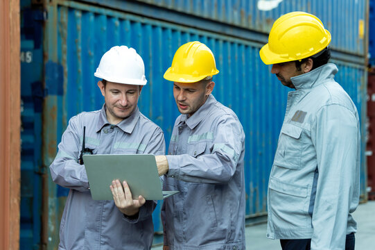 Industrial workers wearing helmets using laptop beside shipping containers, showing logistics, teamwork, safety, technology, inspection, planning, engineering and cargo operations