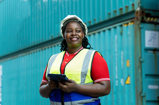 Industrial woman worker wearing helmet and safety vest holding tablet beside shipping containers, showing logistics, technology, inspection, confidence, cargo operations and workplace safety