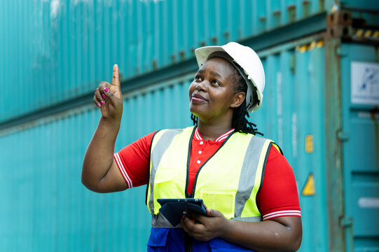 Industrial woman worker wearing helmet and safety vest holding tablet while pointing upward beside shipping containers, showing logistics, inspection, technology, planning and workplace safety