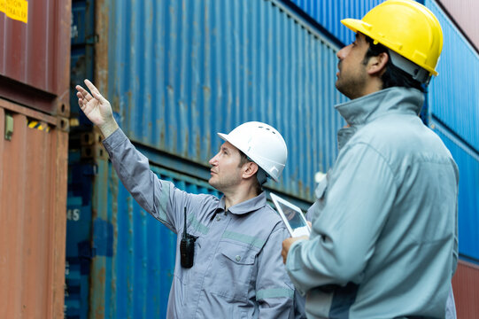 Industrial workers wearing helmets inspecting stacked shipping containers, one man pointing upward while colleague holds tablet, showing logistics, safety, planning and cargo coordination