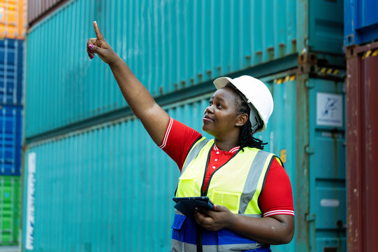 Industrial woman worker wearing helmet and safety vest holding tablet while pointing upward beside shipping containers, showing logistics, inspection, safety, technology and cargo coordination