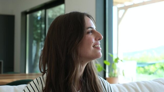 A beautiful young woman smiles softly as she looks out a large window, enjoying a peaceful and contemplative moment in a bright, modern interior.