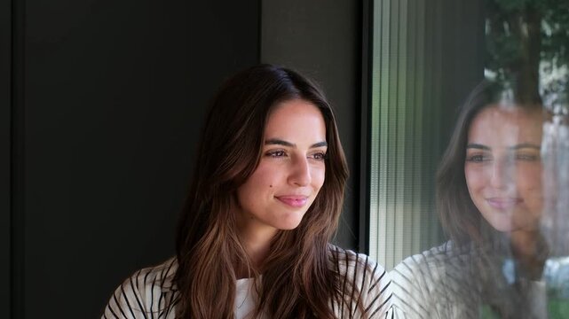 A beautiful young woman smiles brightly as she gazes thoughtfully out of a window. Her serene reflection in the glass conveys happiness and contemplation, highlighting a moment of peace.