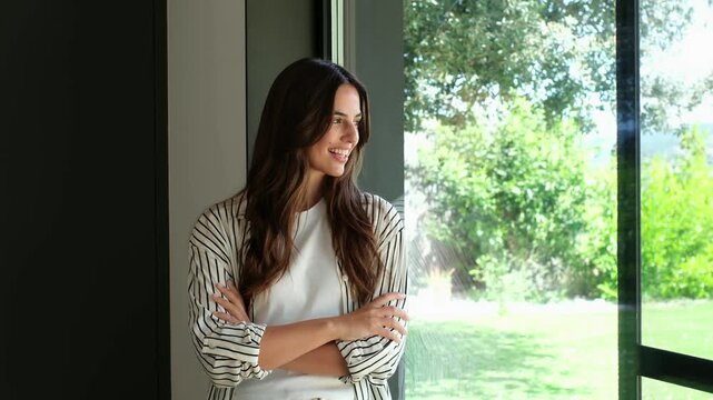 Serene young woman standing by a large window, gazing at the sunny garden outside with a peaceful, content smile, enjoying quiet relaxation in a bright modern interior