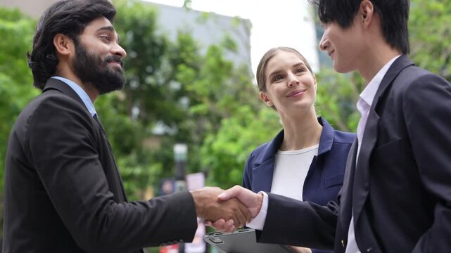 Two businessmen and a businesswoman shake hands and smile at the start of an outdoor business meeting under fresh trees in the public realm of a smart city district